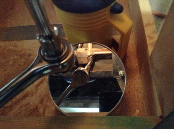 Trunnion bolt head connected to 18" socket set seen through mirror with flashlight while sitting on bucket in front of Mao Shan Table Saw.